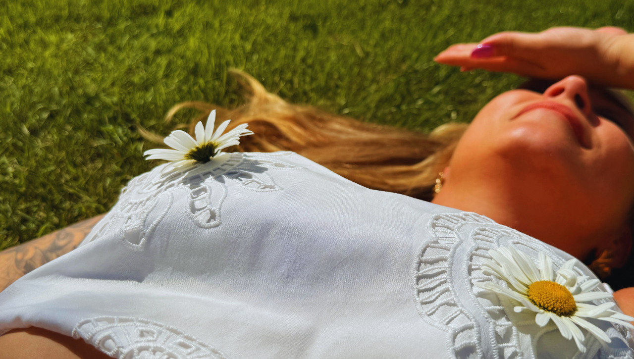 Vrouw ligt in het gras met een witte zomertop over een voorgevormde bh en madeliefjes op haar kleding.