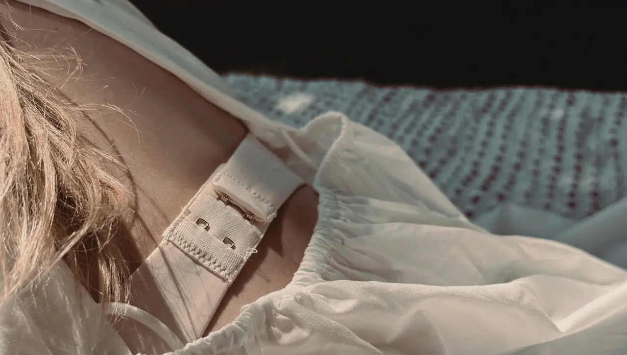 Close-up van een bh-bandje met verstelbare sluiting op de schouder, onder een loshangende witte blouse in zacht ochtendlicht. Beeld bij wat maakt een bh ondersteunend.